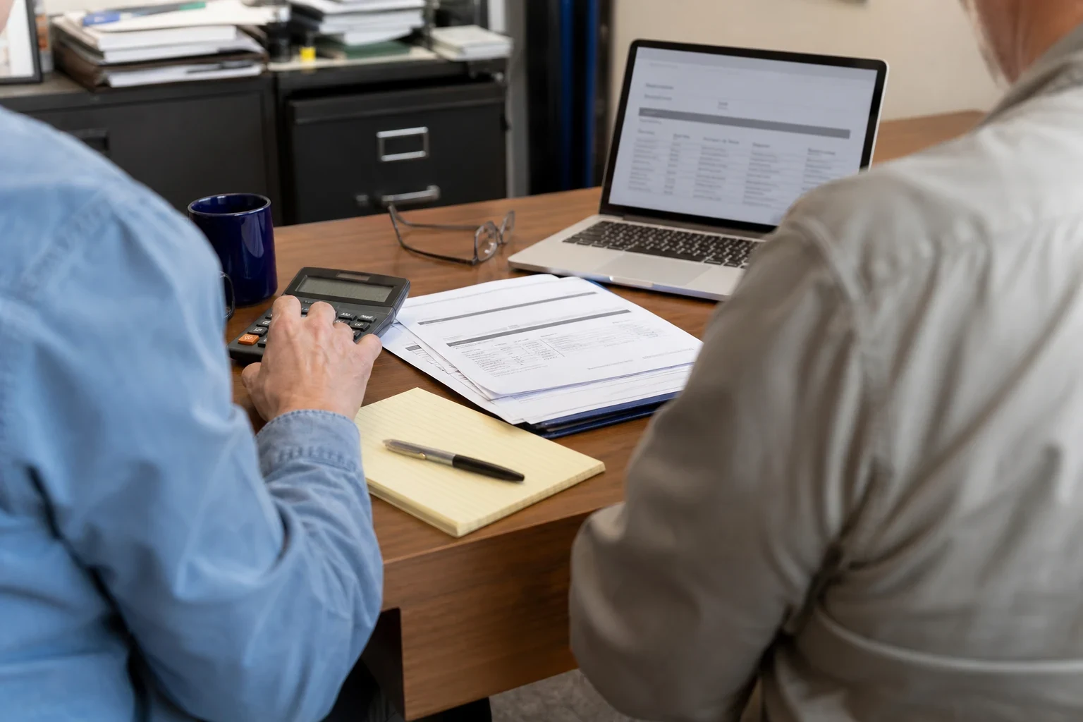 Over-the-shoulder view of two people at an office desk, one using a calculator beside printed statements, a yellow notepad with pen, and an open laptop.